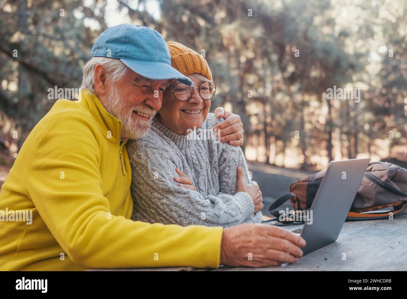 Porträt, Nahaufnahme eines niedlichen Paares von älteren Menschen mittleren Alters, die im Freien einen Computer benutzen und an einem Holztisch im Wald der Berge in der Natur sitzen, mit Bäumen um sie herum. Stockfoto