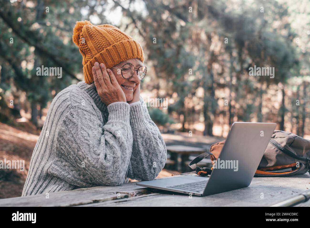Porträt, Nahaufnahme einer niedlichen Person des alten mittleren Alters, die im Freien einen Computer benutzt und an einem Holztisch im Wald des Berges in der Natur sitzt, mit Bäumen um sie herum. Stockfoto