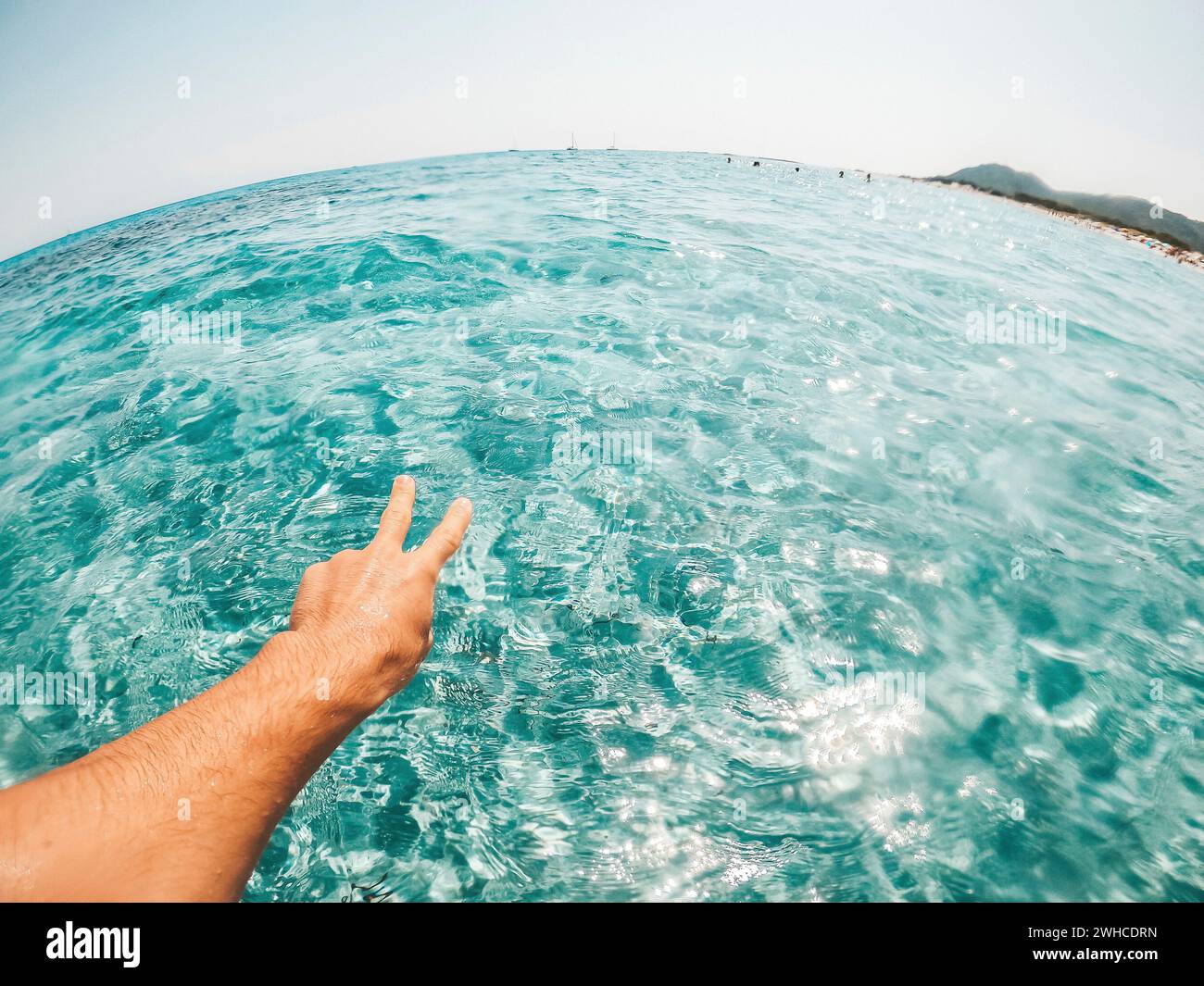 Pov und erste Ansicht der Person, die im Wasser des Strandes im Meer im blau-türkisfarbenen und tropischen Wasser schwimmend das Sieg- und Freiheitszeichen mit seiner Hand tut Stockfoto
