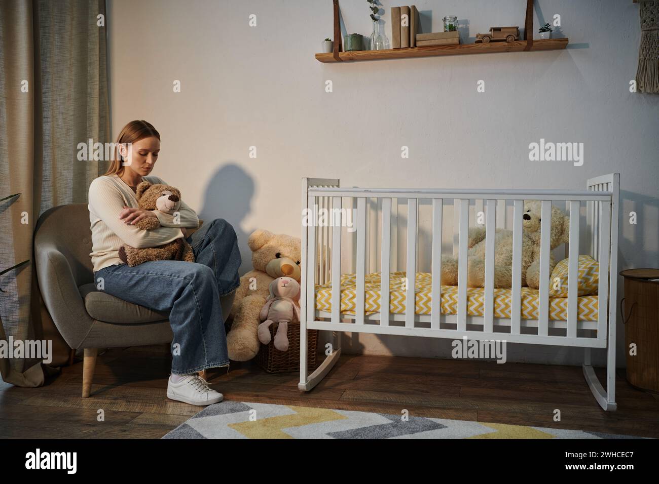 Verzweifelte junge Frau mit weichem Spielzeug, die im Sessel neben dem Kinderbett sitzt, in einem dunklen Kinderzimmer zu Hause Stockfoto