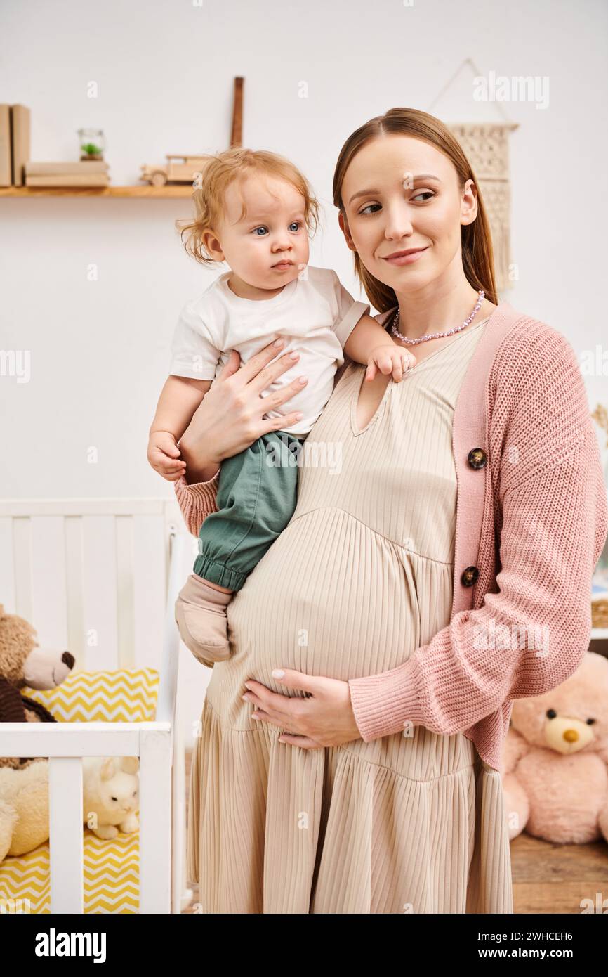 Glückliche junge schwangere Frau, die mit dem Kleinkind im Kinderzimmer zu Hause steht, glückliche Mutterschaft Stockfoto