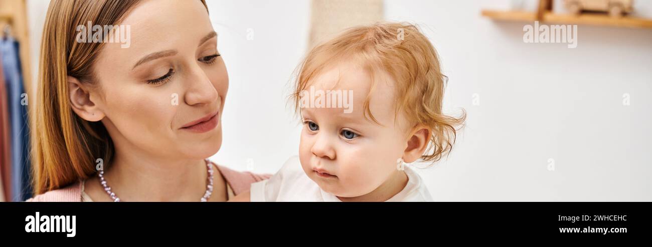 Fröhliche attraktive Frau, die einen kleinen Kleinkinder in den Händen hält, im Kinderzimmer, horizontales Banner Stockfoto