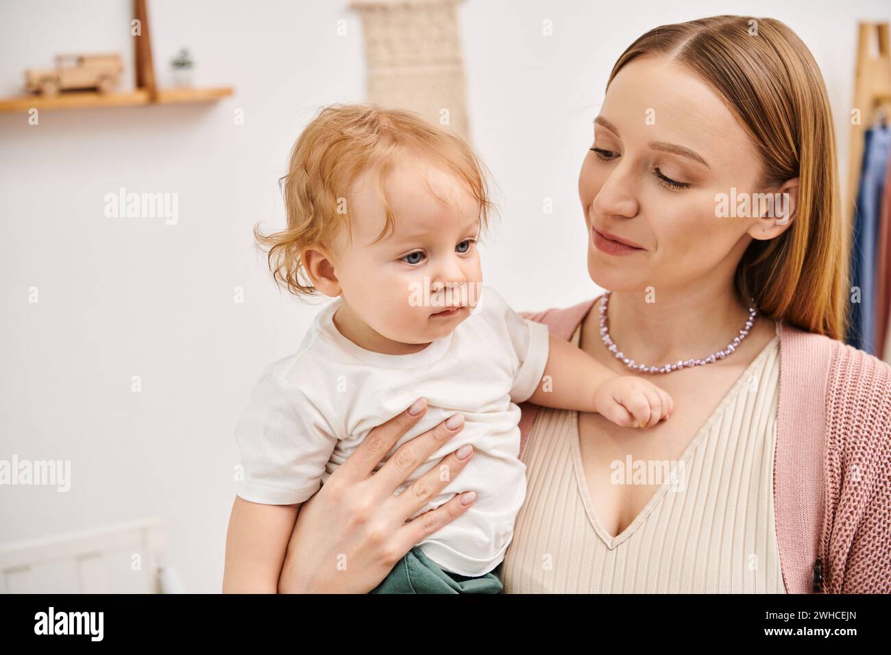 Fröhliche, attraktive Frau, die einen kleinen Kleinkinder in den Händen hält, im Kinderzimmer, glückliche Mutterschaft Stockfoto