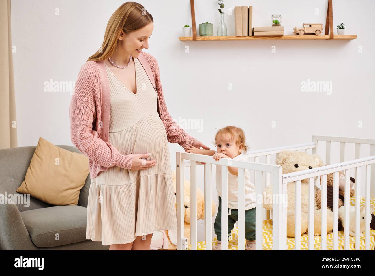 Lächelnde schwangere Frau, die einen kleinen Sohn umschließt, der in einem Kinderzimmer steht, moderne Erziehung Stockfoto