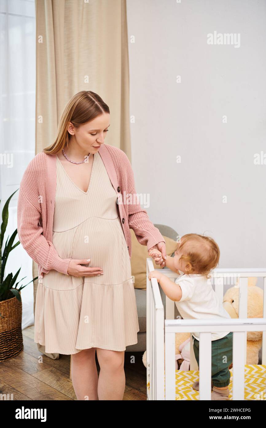 Glückliche schwangere Frau, die Hand eines kleinen Sohnes hält, der in der Krippe im gemütlichen Kinderzimmer steht, Mutterschaft Stockfoto