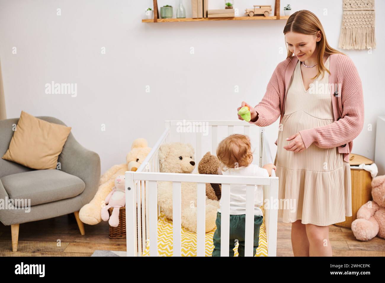 Fröhliche schwangere Frau, die Spielzeug hält und mit dem Kleinkind-Sohn spielt, der im Kinderzimmer in der Krippe steht Stockfoto
