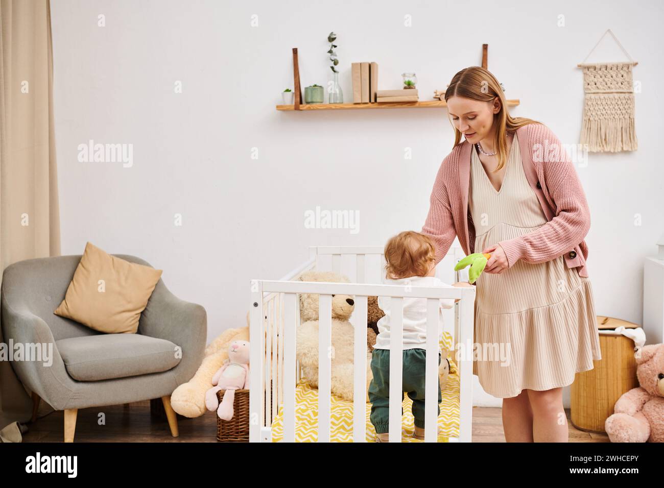 Lächelnde schwangere Frau, die Spielzeug hält und mit dem Kleinkindsohn spielt, der im Kinderzimmer in der Krippe steht Stockfoto