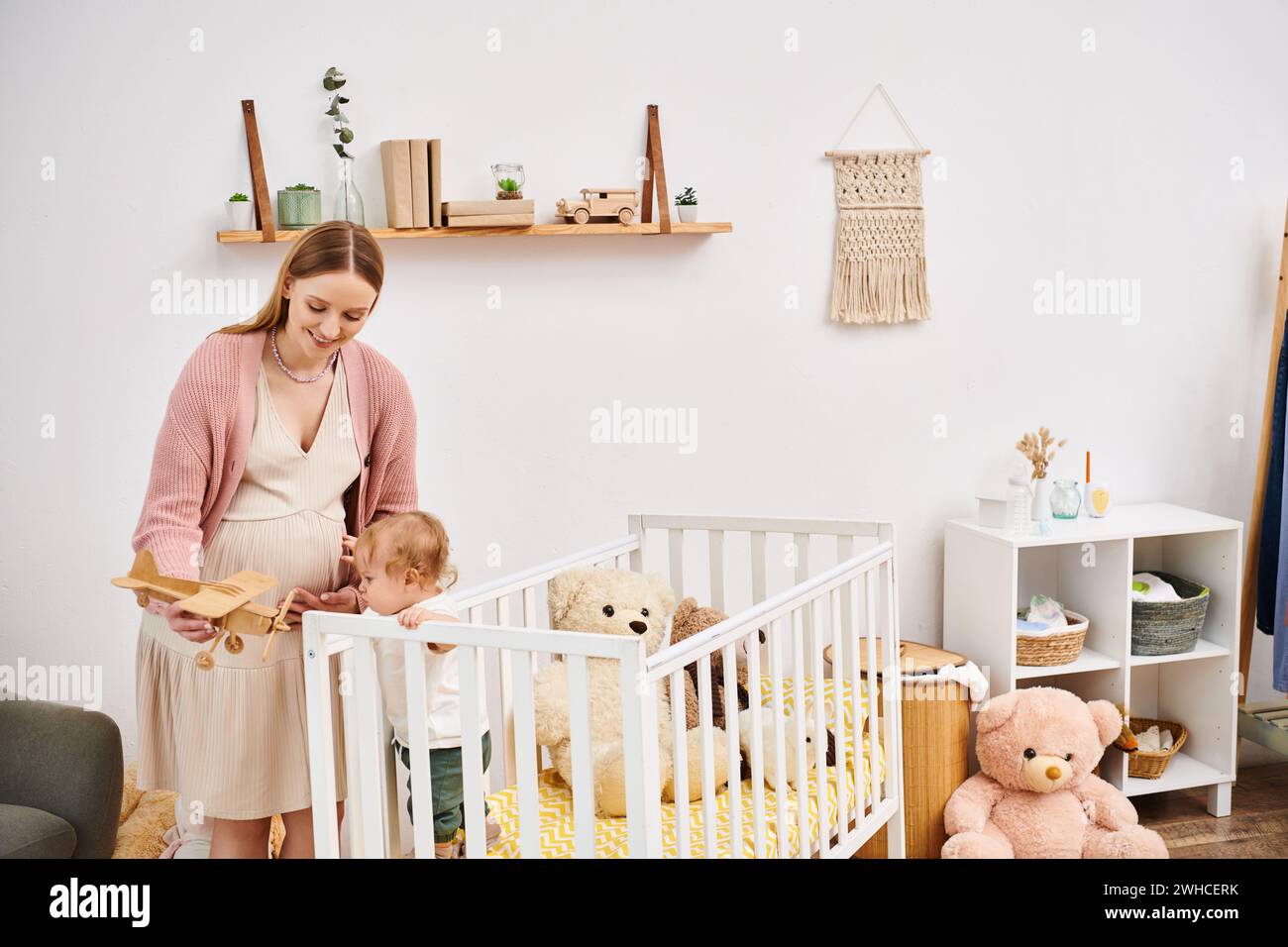 Lächelnde schwangere Frau, die mit Spielzeugflugzeug spielt und Kleinkind-Junge im Kinderzimmer zu Hause, Mutterschaft Stockfoto