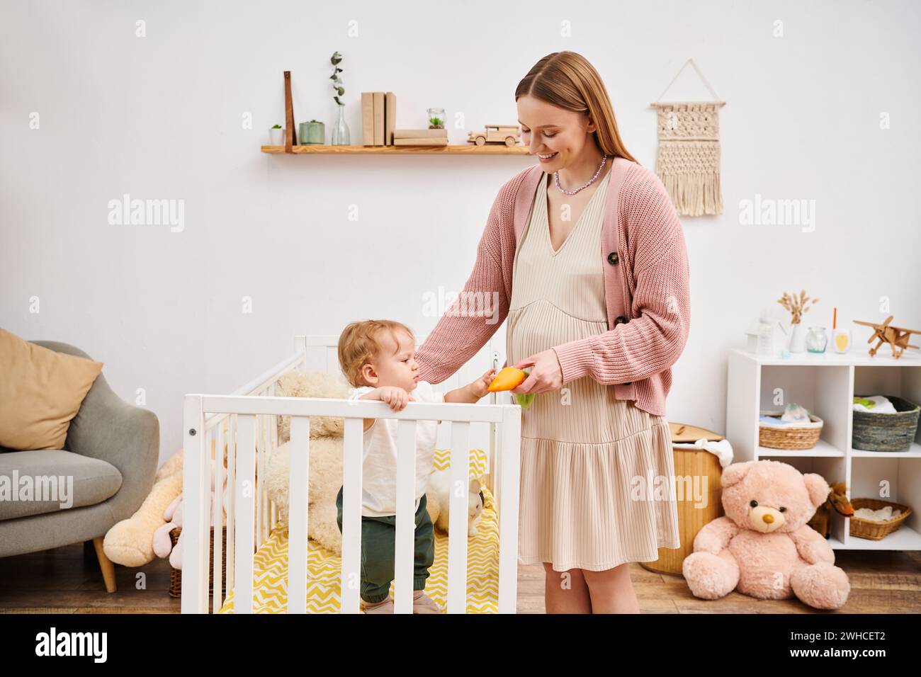 Fröhliche schwangere Frau, die Spielzeug hält, während sie mit dem kleinen Sohn spielt, der im Kinderzimmer im Kinderbett steht Stockfoto