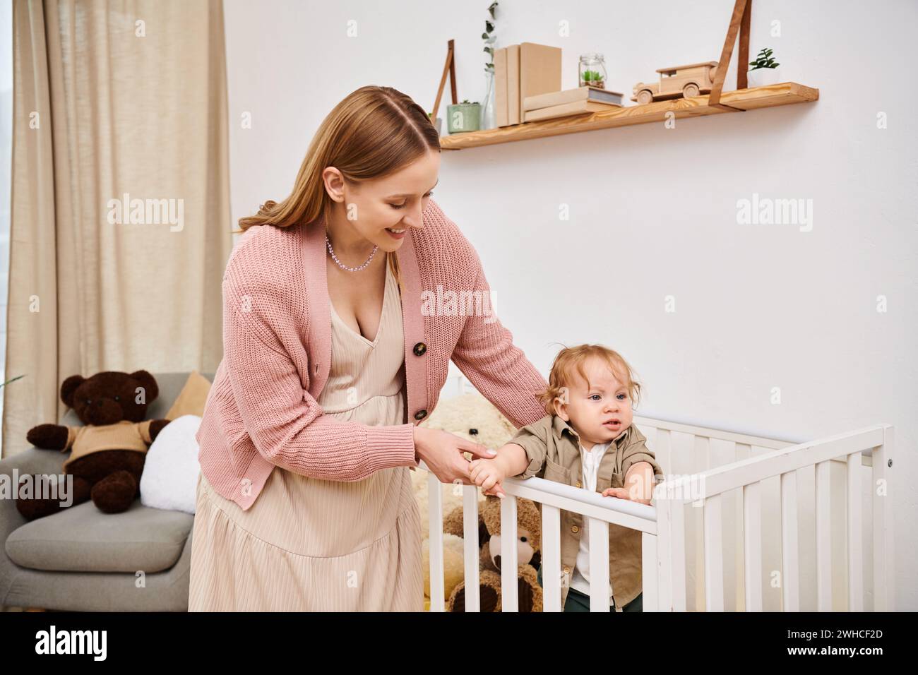 Fröhliche Frau, die den Kleinkind-Sohn unterstützt, der im Kinderzimmer zu Hause im Kinderbett steht, glückselige Mutterschaft Stockfoto