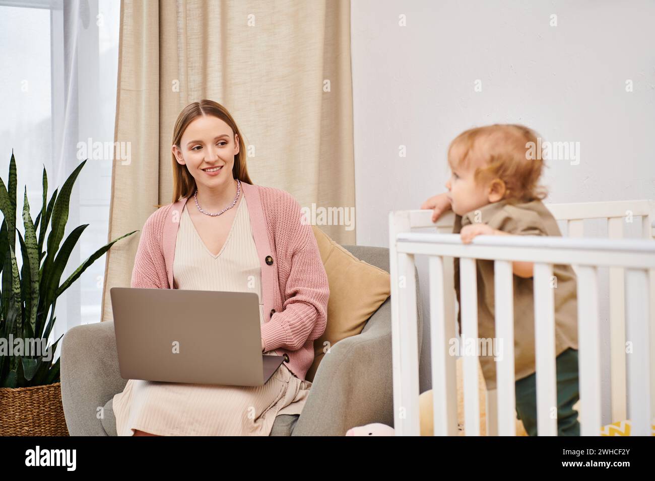 Glückliche Mutter, die an einem Laptop arbeitet, in der Nähe eines niedlichen Kindes im Kinderzimmer, Multitasking-Frau Stockfoto