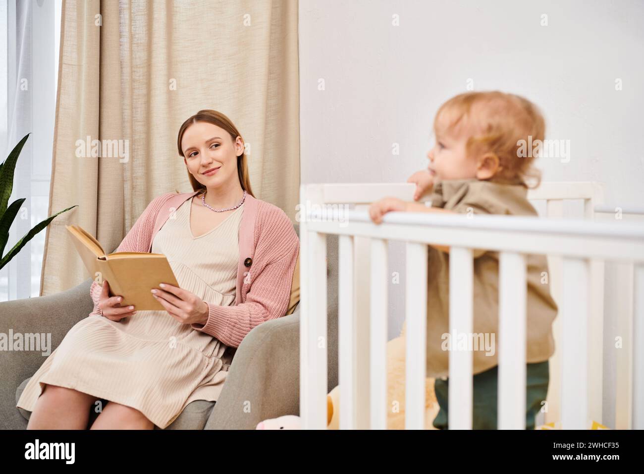 Glückliche schwangere Frau, die ein Buch in der Nähe des entzückenden Kleinkindsohnes im Kinderzimmer zu Hause liest, Mutterschaft Stockfoto