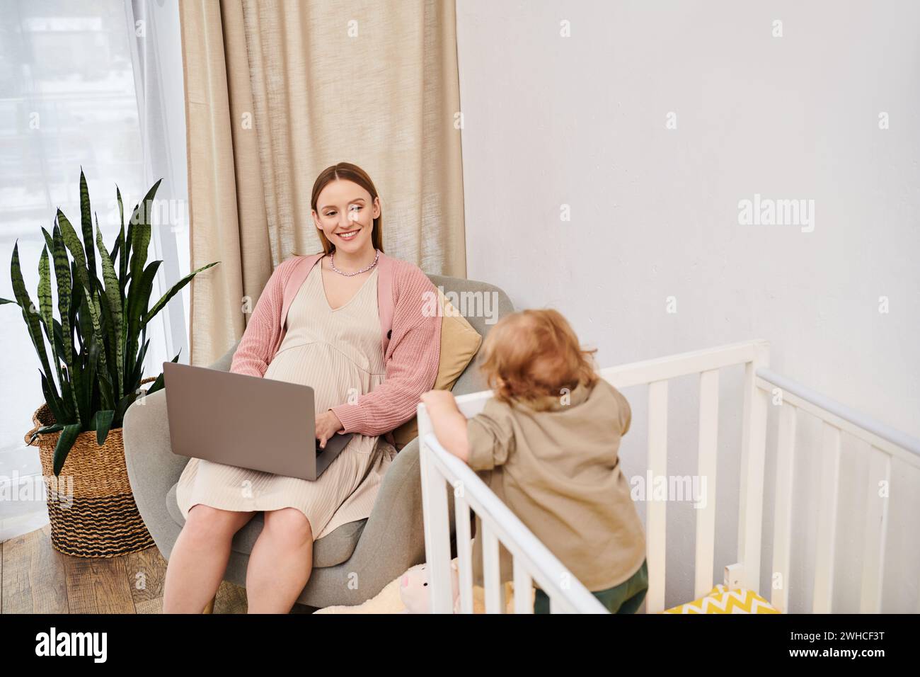 Lächelnde Frau, die an einem Laptop in der Nähe des Kleinkindes arbeitet, in der Babywanne im Kinderzimmer, Multitasking Stockfoto