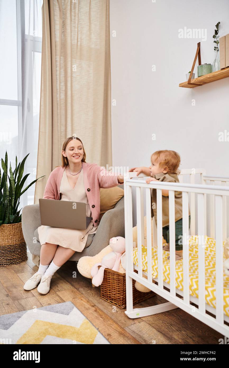 Fröhliche Frau, die an Laptop in der Nähe des niedlichen Kindes im Kinderzimmer arbeitet, Multitasking-Mutter Stockfoto