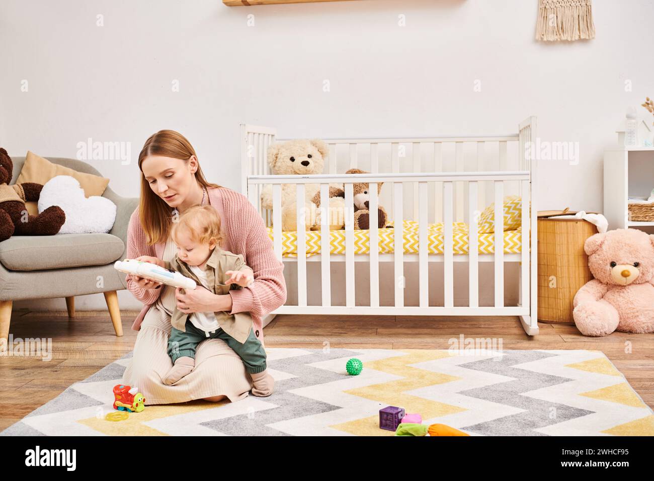 Junge Mutter, die mit dem niedlichen Kleinkind-Sohn in der Nähe eines Kinderbetts auf dem Boden im Kinderzimmer spielt, glückliche Mutterschaft Stockfoto