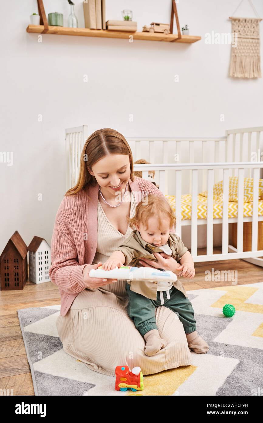 Glückliche Mutter, die mit einem süßen Kleinkind in der Nähe des Kinderbetts spielt, auf dem Boden im Kinderzimmer, glückselige Mutterschaft Stockfoto