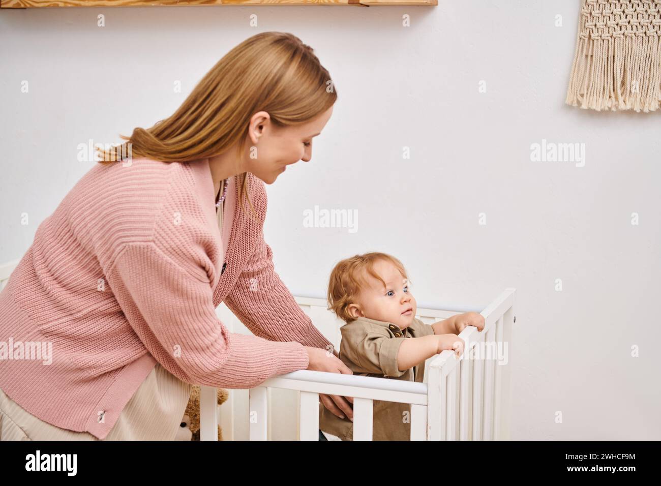 Lächelnde Frau, die den Sohn eines Kleinkindes unterstützt, der im Kinderzimmer zu Hause steht, glückliche Mutterschaft Stockfoto