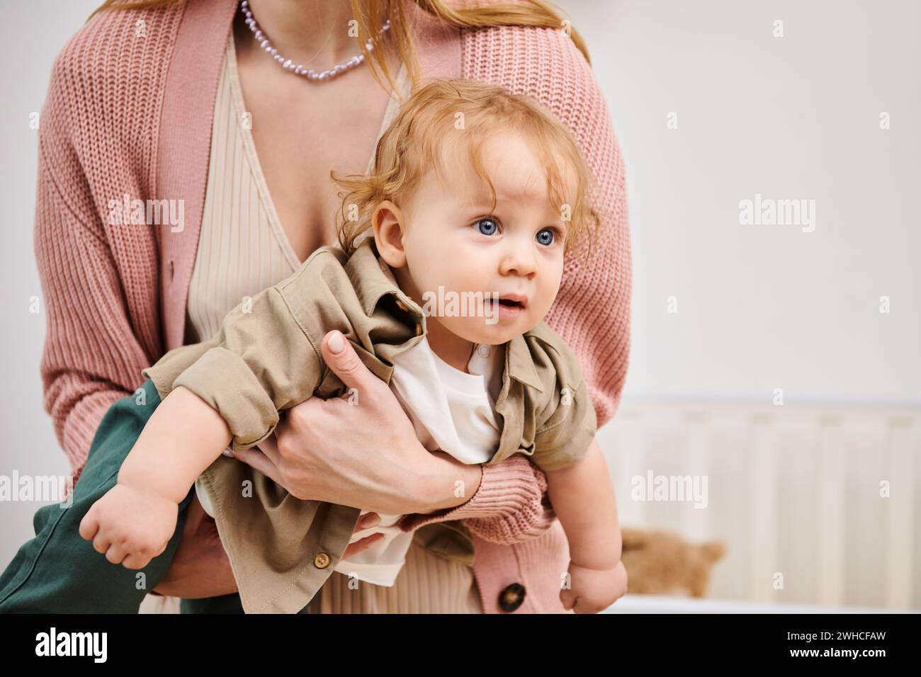 Entzückender Junge, der in den Händen seiner Mutter im Kinderzimmer zu Hause wegschaut, glückliche Kindheit Stockfoto