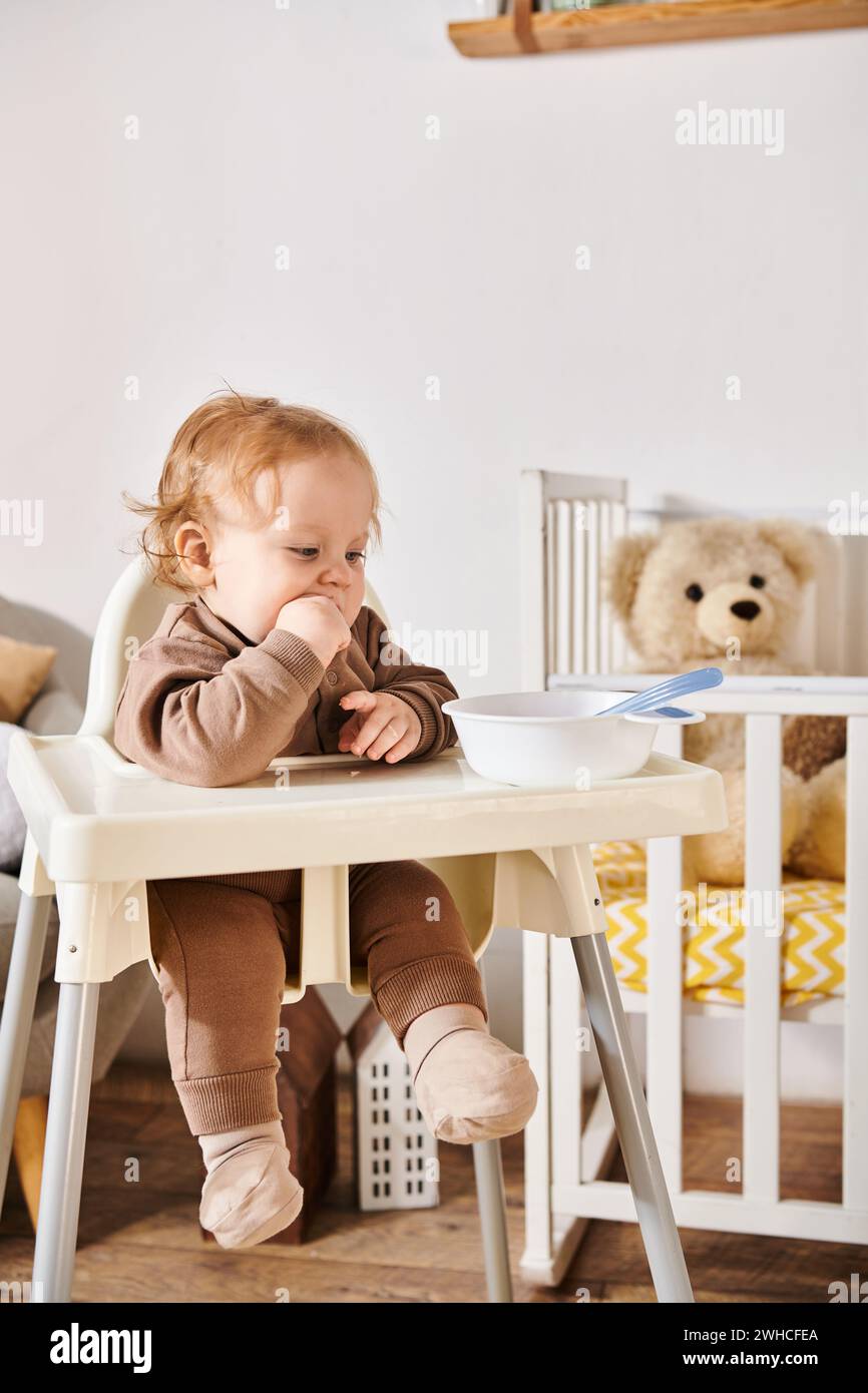 Niedliches kleines Kind, das im Kinderstuhl neben der Schüssel sitzt und im Kinderzimmer frühstückt, glückselige Kindheit Stockfoto