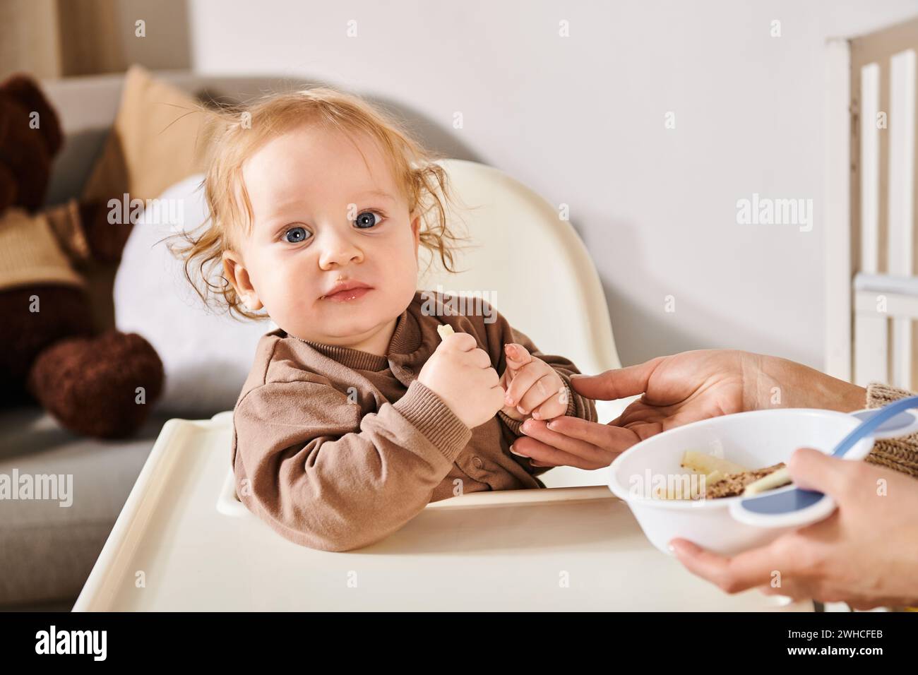 Entzückender Junge, der im Kinderstuhl sitzt und in die Kamera neben der Mutter blickt, mit einer Schüssel Frühstück Stockfoto