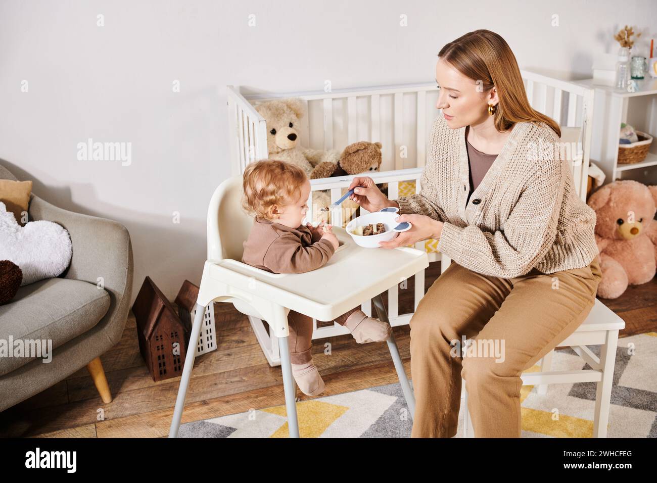 Liebevolle Frau, die den Jungen mit Frühstück auf dem Kinderstuhl im Kinderzimmer füttert, glückselige Mutterschaft Stockfoto