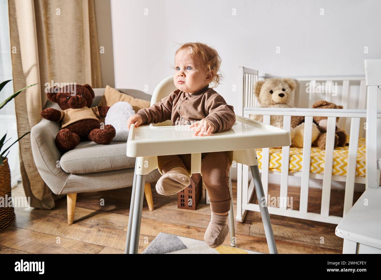 Kleiner Junge sitzt im Kinderstuhl und blickt weg in einem gemütlichen Kinderzimmer mit Kinderbett und Teddybären Stockfoto