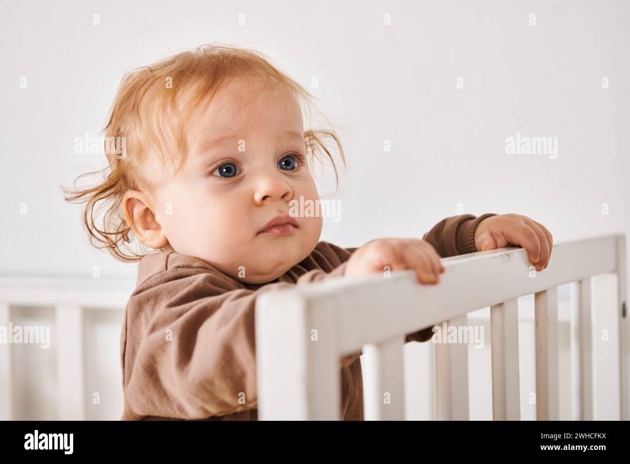 Porträt des liebenswerten Kindes, das in der Krippe im Kinderzimmer steht und wegschaut, glückliche Babyhood Stockfoto