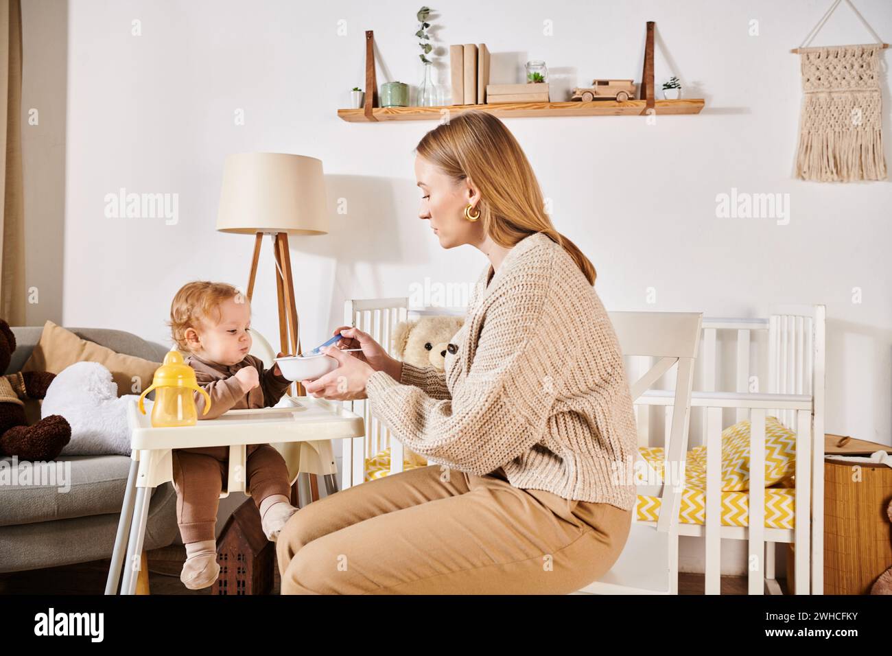 Junge Mutter füttert kleinen Sohn mit Frühstück auf Kinderstuhl im Kinderzimmer, moderne Erziehung Stockfoto