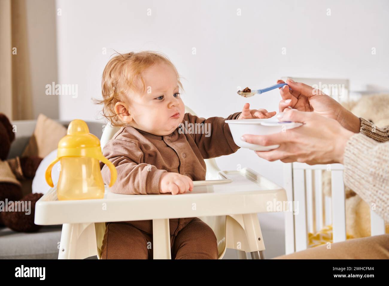 Mutter füttert entzückenden Sohn mit Frühstück auf Kinderstuhl im Kinderzimmer, moderne Erziehung Stockfoto