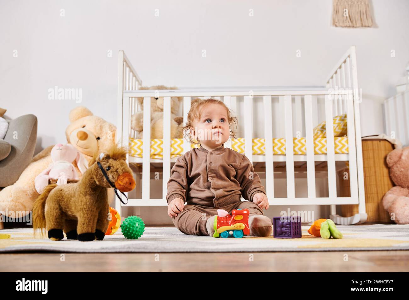 Kleiner unschuldiger Junge, der auf dem Boden neben dem Kinderbett und den Kuschelspielzeugen im gemütlichen Kinderzimmer sitzt, glückliche Babynahrung Stockfoto