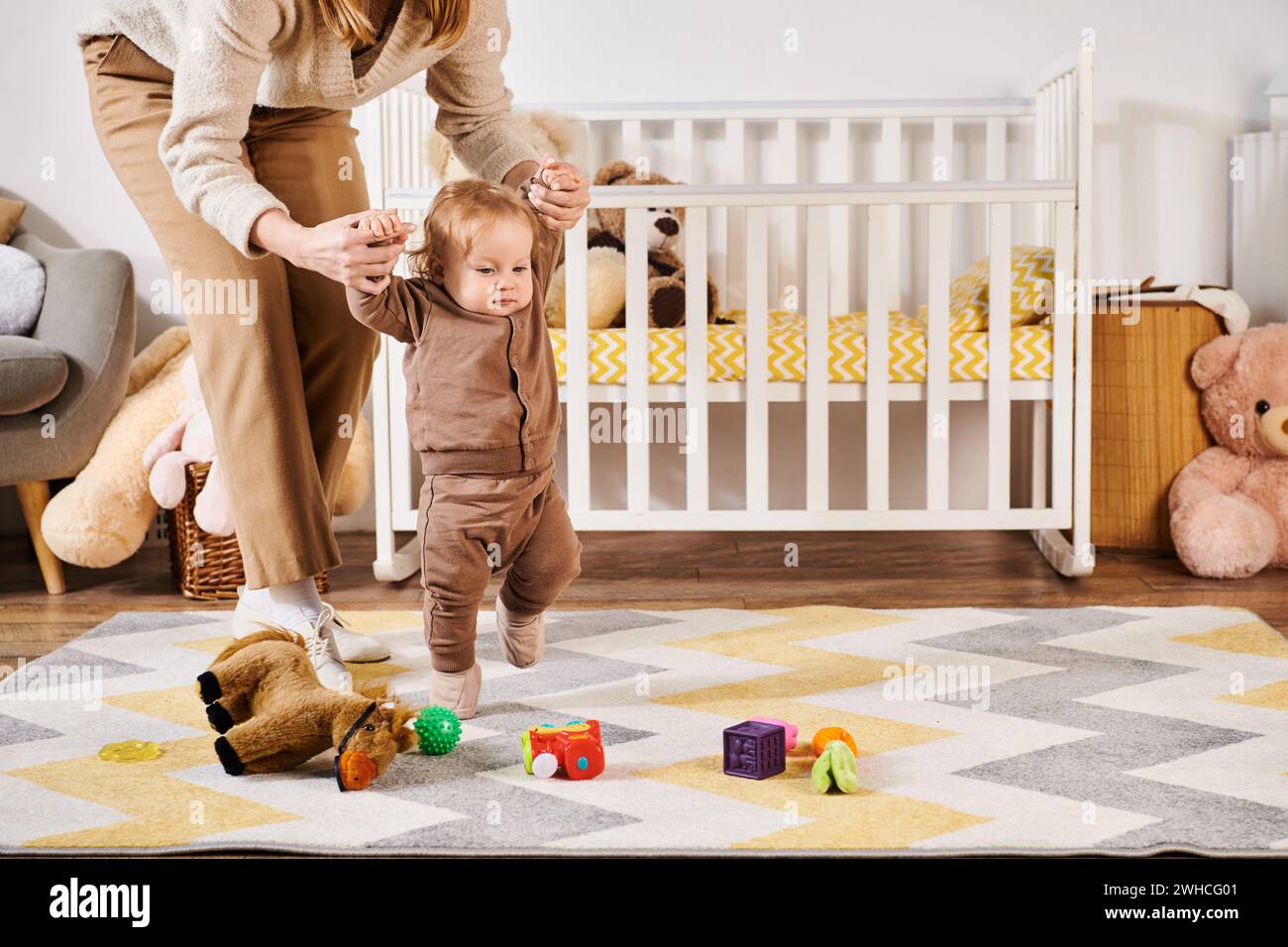 Junge Mutter, die Hände eines kleinen Sohnes hält, der in der Nähe von Spielzeug und Krippe im Kinderzimmer läuft, Unterstützung Stockfoto
