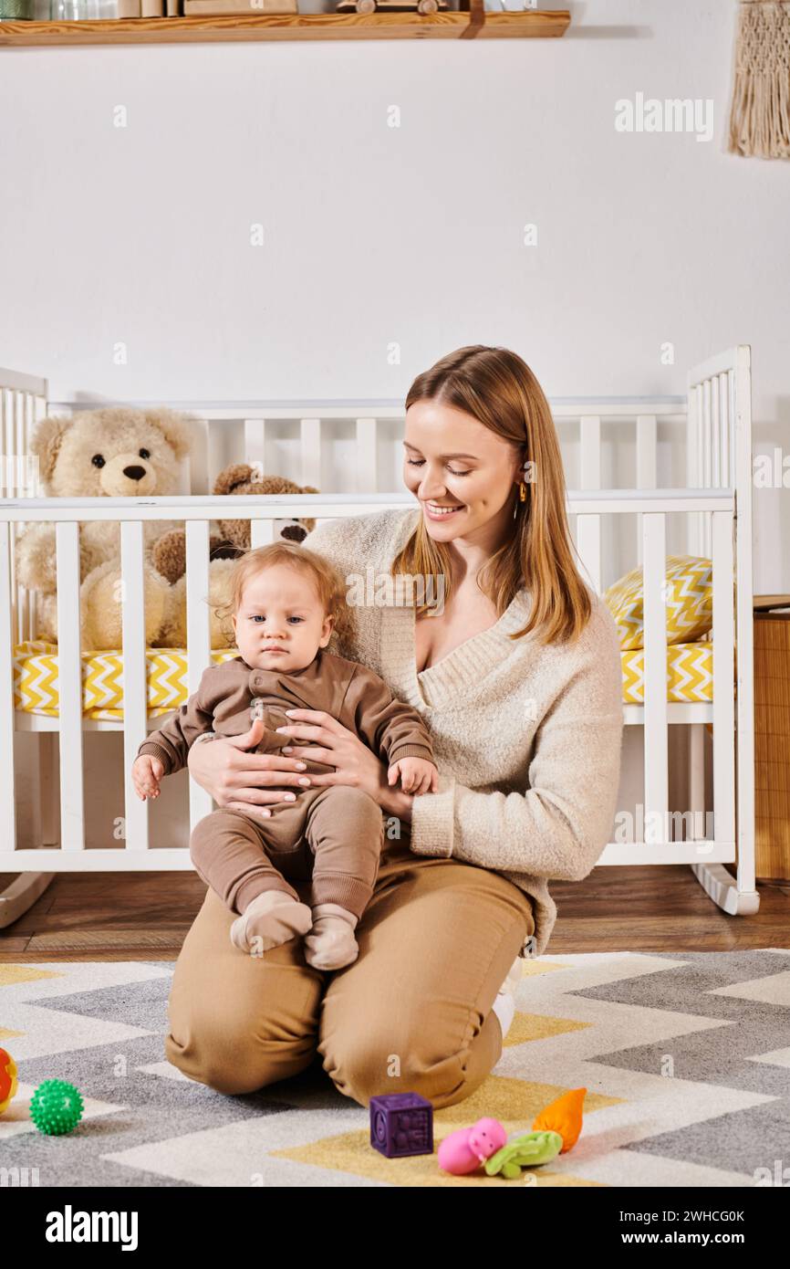 Lächelnde Mutter, die den Kleinkinder in der Nähe von Spielzeugen auf dem Boden im gemütlichen Kinderzimmer umschließt, glückliche Mutterschaft Stockfoto