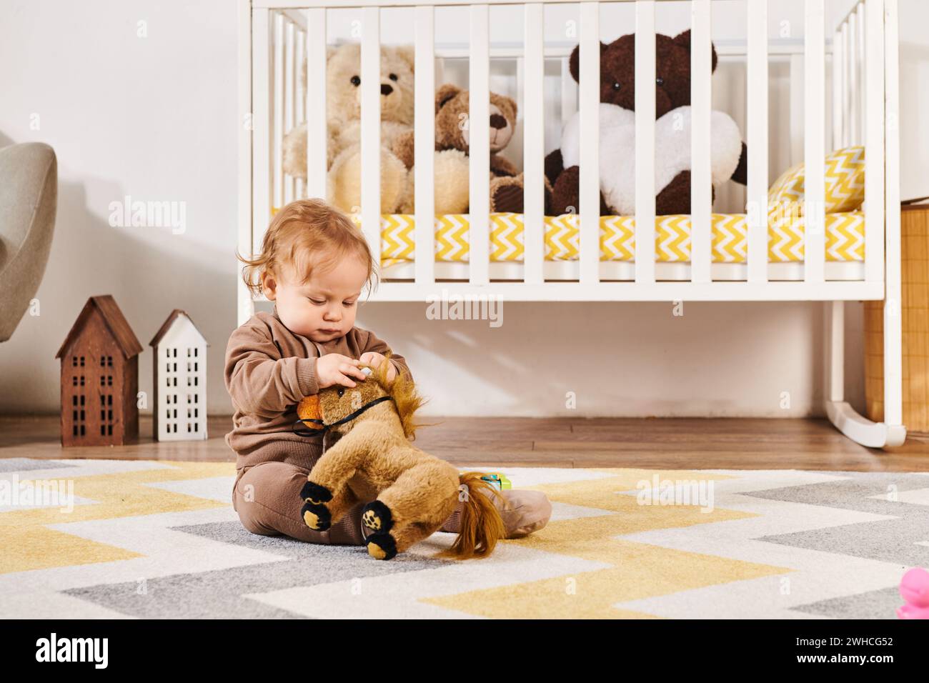 Niedlicher kleiner Junge, der auf dem Boden sitzt und mit einem Spielzeugpferd in der Nähe des Kinderbetts in einem gemütlichen Kinderzimmer spielt Stockfoto