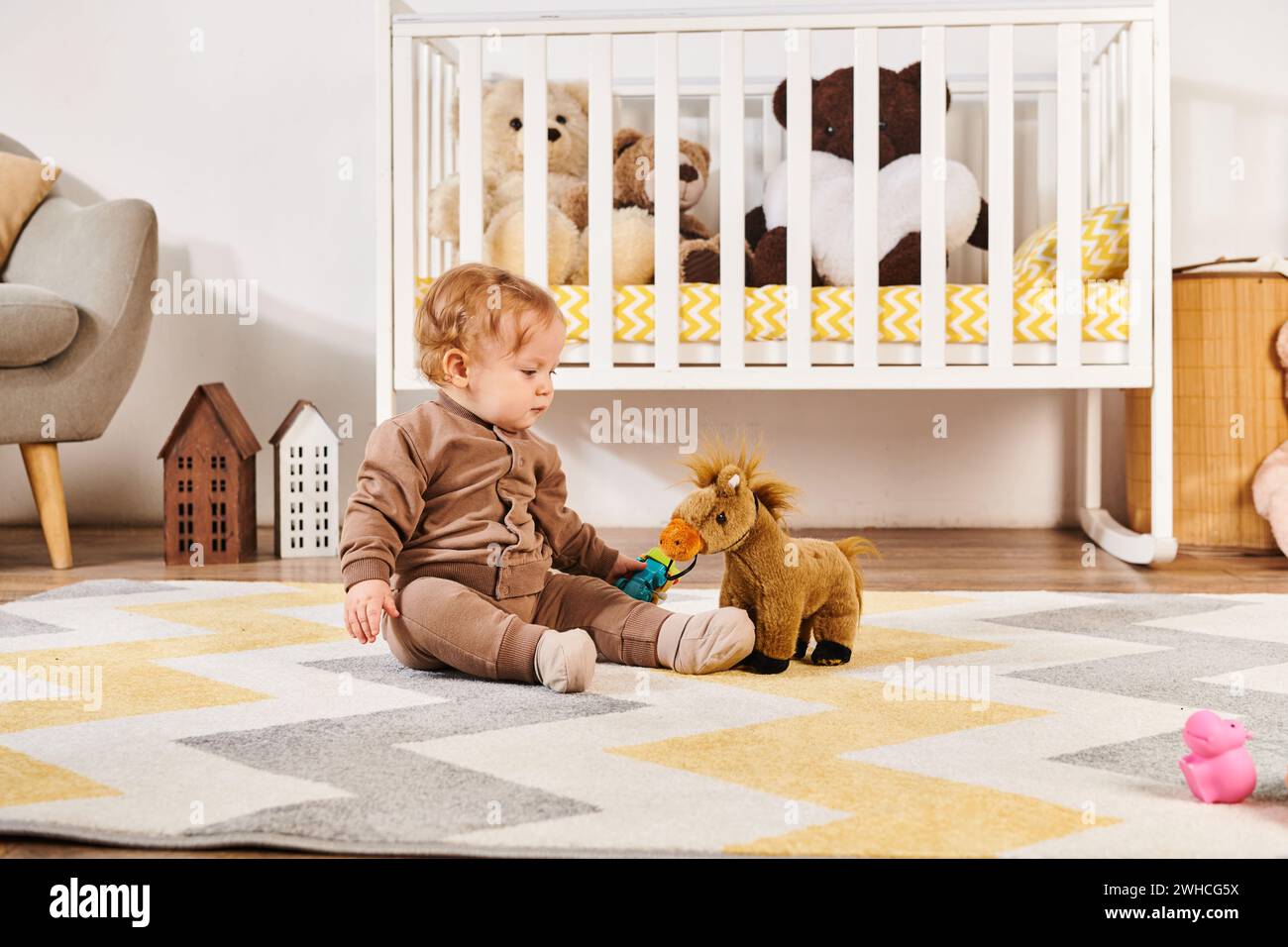 Entzückender Kleinkind, der auf dem Boden sitzt und mit einem Spielzeugpferd in der Nähe des Kinderzimmers spielt Stockfoto