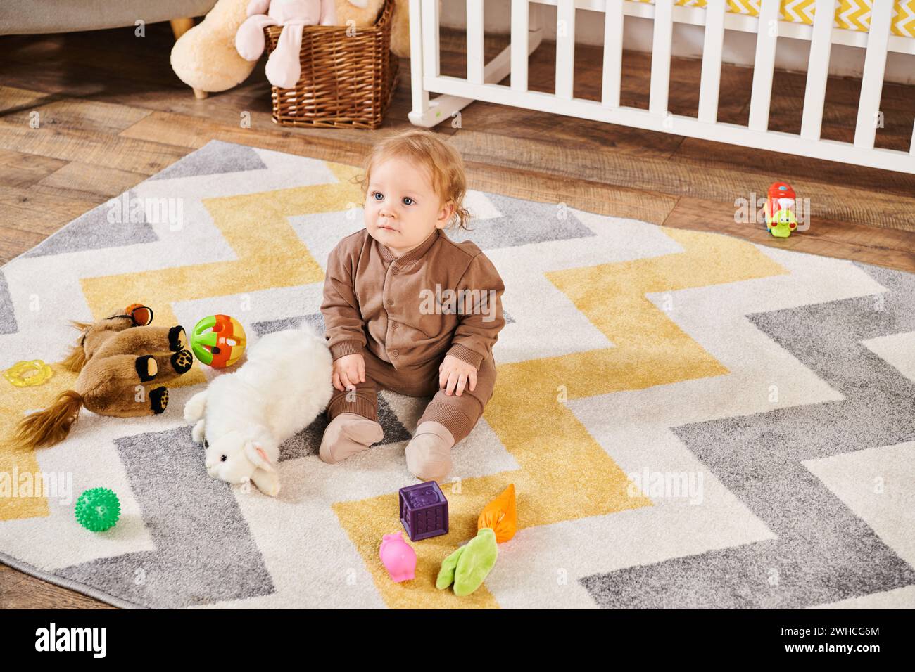 Entzückendes Kind, das auf dem Boden neben Kuschelspielzeug und Kinderbett im gemütlichen Kinderzimmer sitzt, fröhliches Kleinkind Stockfoto