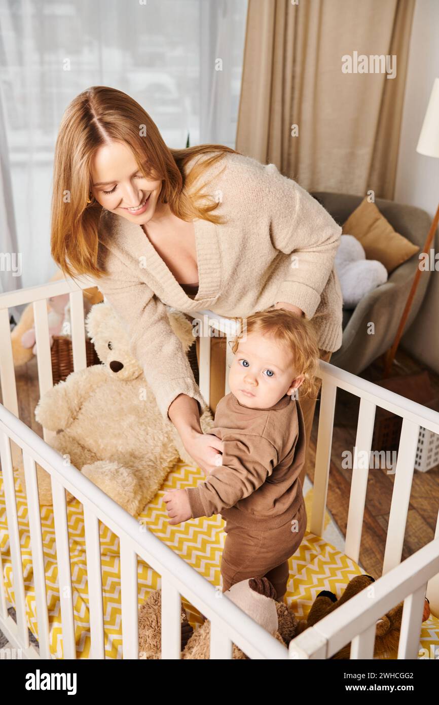 Junge, fröhliche Frau lächelt in der Nähe eines kleinen Kindes, das in der Krippe im Kinderzimmer zu Hause steht, Mutterschaft Stockfoto