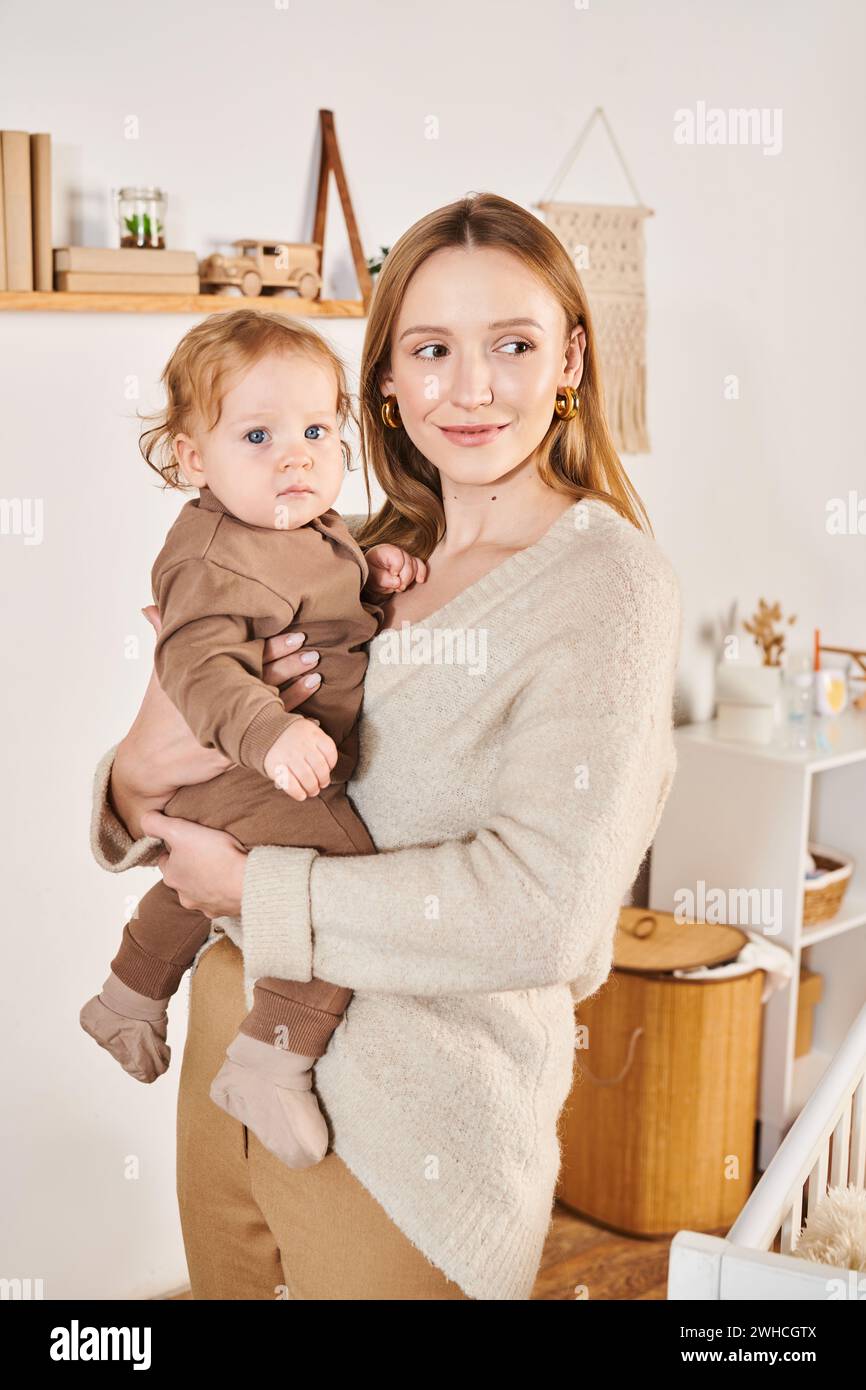 Fröhliche attraktive Frau mit süßem kleinen Sohn in Händen, die im Kinderzimmer steht, moderne Mutterschaft Stockfoto
