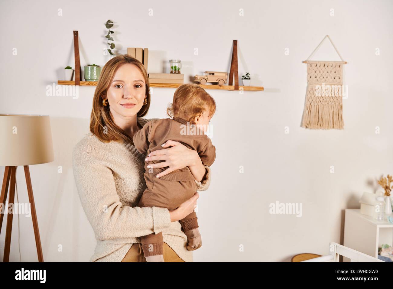Glückliche junge Frau, die einen kleinen Kleinkinder im Kinderzimmer hält und umschließt, glückselige Mutterschaft Stockfoto