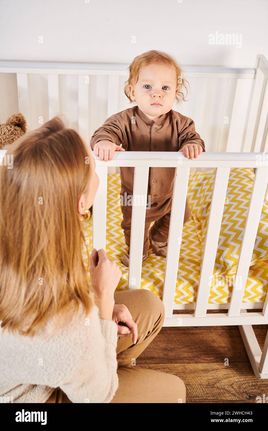 Frau, die den süßen Kleinkinder ansieht, der im Kinderzimmer zu Hause im Kinderbett steht, glückliche Mutterschaft Stockfoto