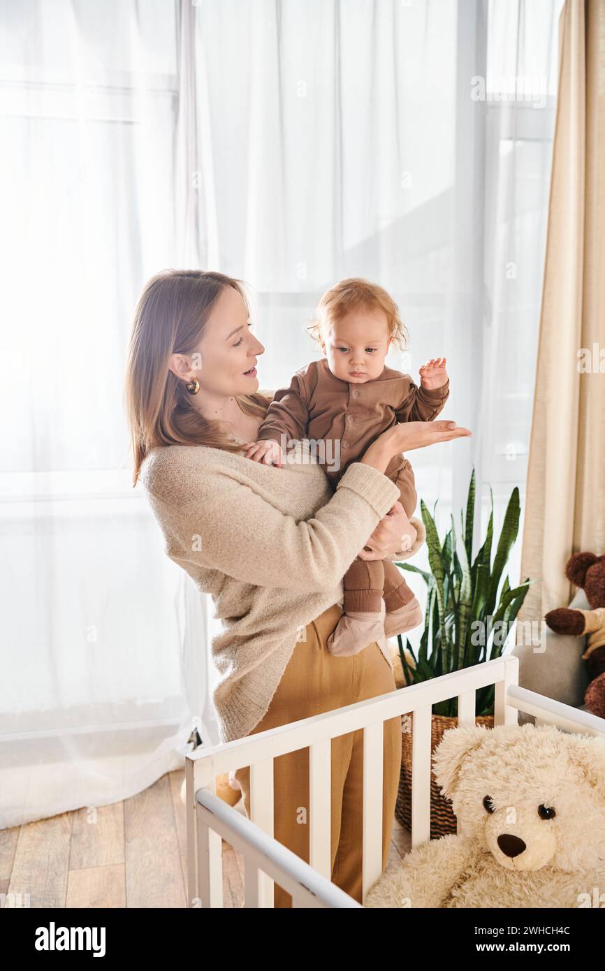 Fröhliche Frau, die den kleinen Sohn in der Hand in der Nähe von Krippe im Kinderzimmer zu Hause hält, glückselige Mutterschaft Stockfoto