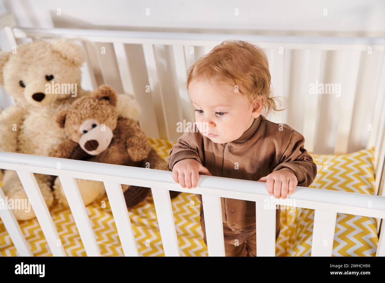 Hochwinkelansicht des kleinen Jungen im Strampler, der im Kinderbett in der Nähe von Kuschelspielzeug im Kinderzimmer steht Stockfoto