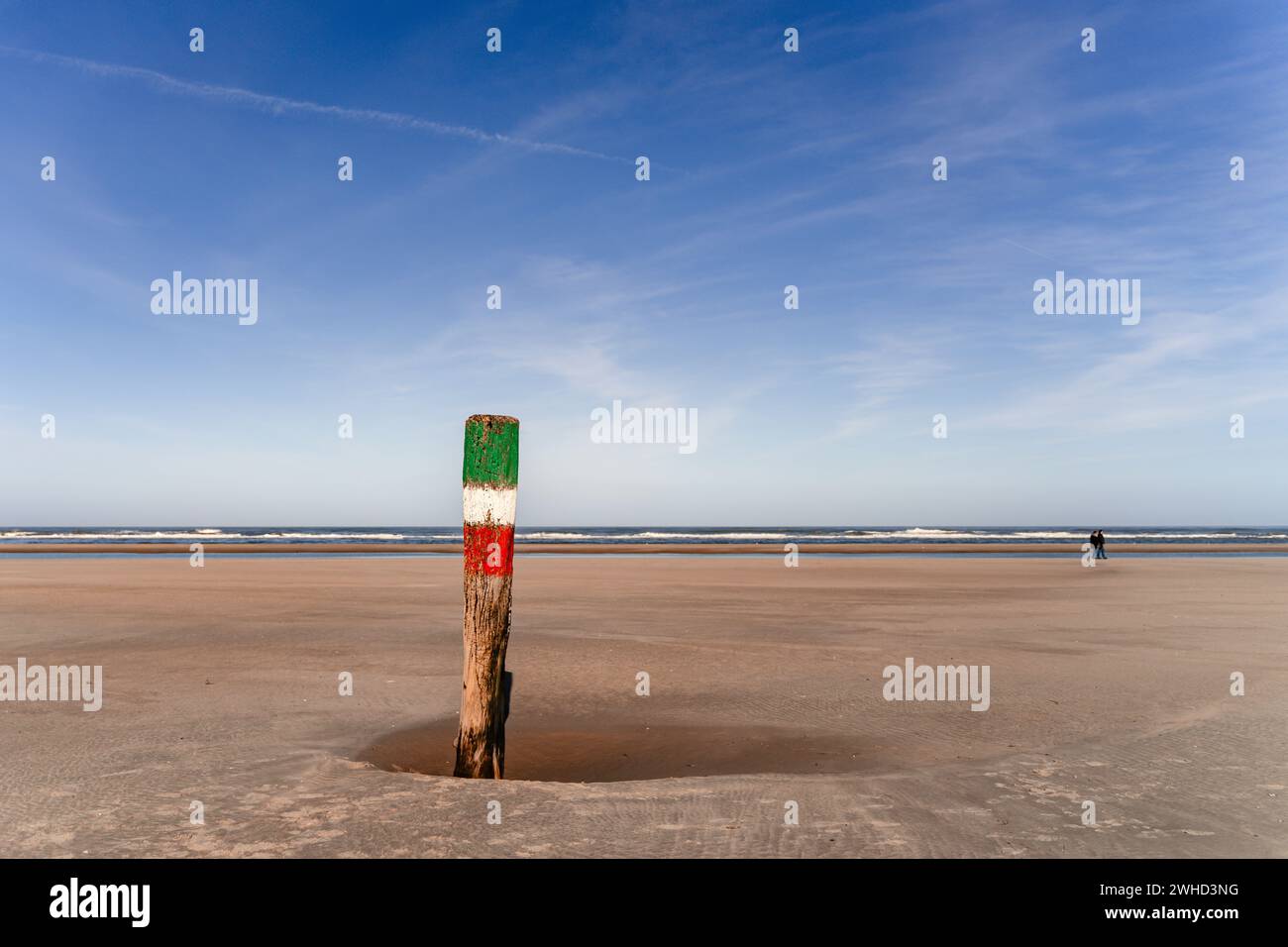 Holzpfahl im Sand am Strand der Insel Norderney an einem sonnigen Tag, zwei verschwommene Menschen im Hintergrund Stockfoto