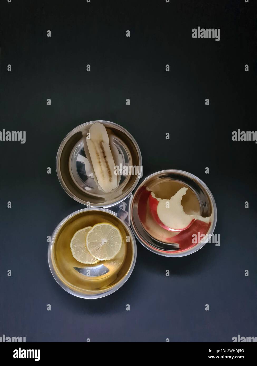 Gebissene Apfelscheibe in Scheiben geschnittene BananenZitronenscheibe in einer der drei silbernen Schalen vor einem grauen Diätthema Stockfoto