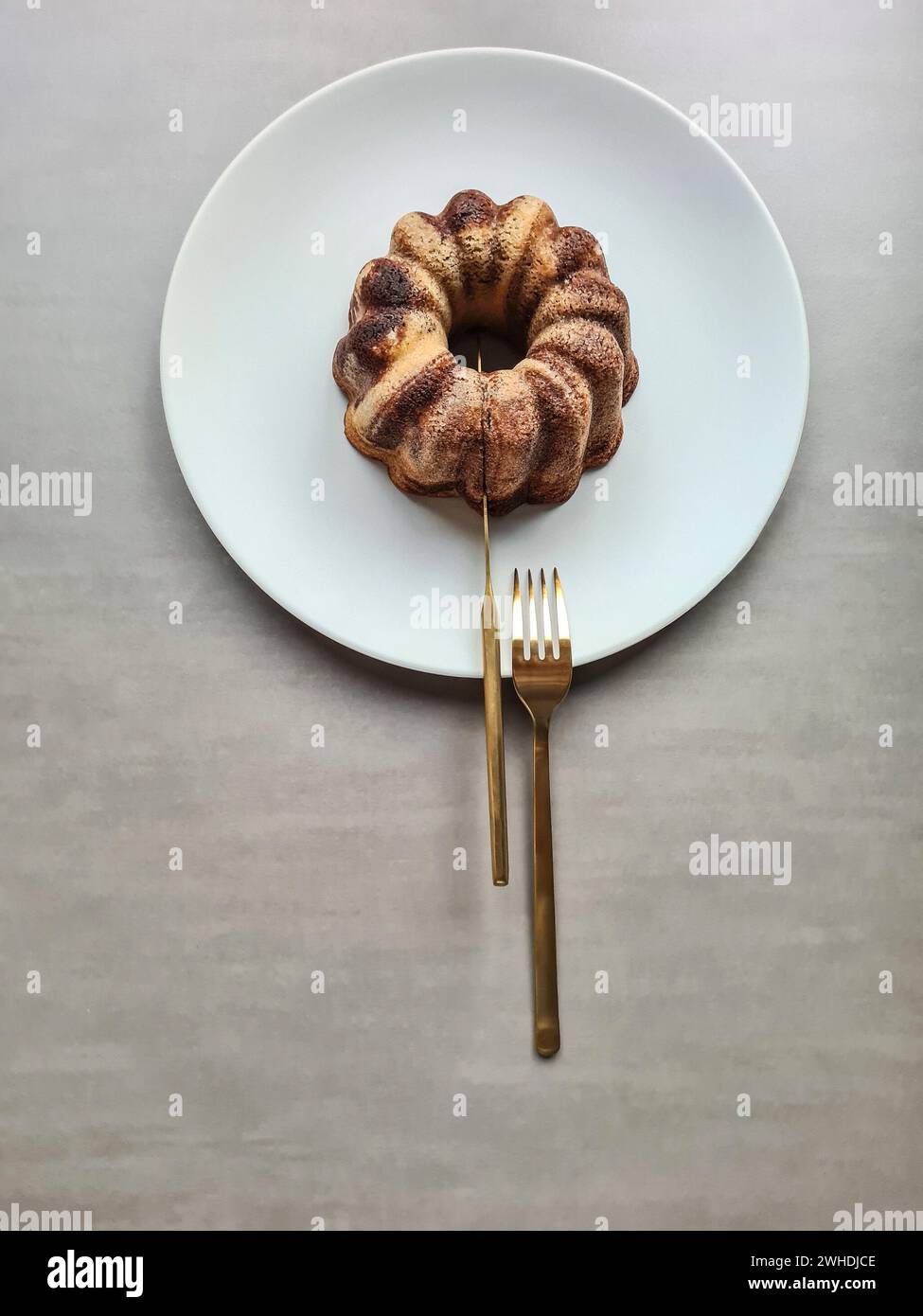 Ein goldfarbenes Messer im Schnitt des Marmortortes auf einem hellgrauen Teller mit Gabel als modernes Lebensmittelstillleben Stockfoto