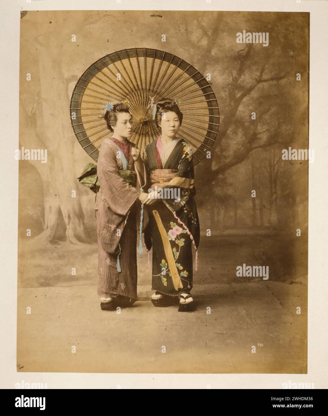 Zwei japanische Frauen im Kimono, durchgehendes Studioporträt mit Hintergrund, leicht nach rechts gerichtet, mit großem Sonnenschirm stehend und Hände haltend....Japan handkoloriert mit Aquarell-Fotografie um 1870er Jahre Stockfoto