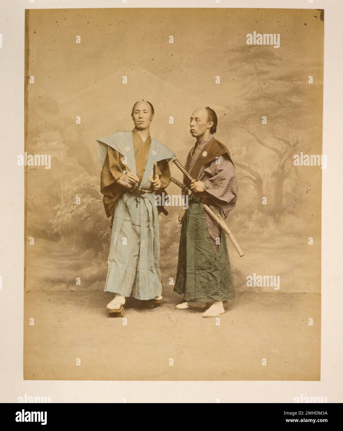 Zwei japanische Männer, als Samurai gekleidet, durchgehendes Studioporträt mit Hintergrund, stehend, einer nach vorne und der andere nach links gerichtet, jeweils mit zwei Schwertern ca. 1877 Yokohama....Japan handkoloriert mit Aquarell-Fotografie um 1870er Jahre Stockfoto