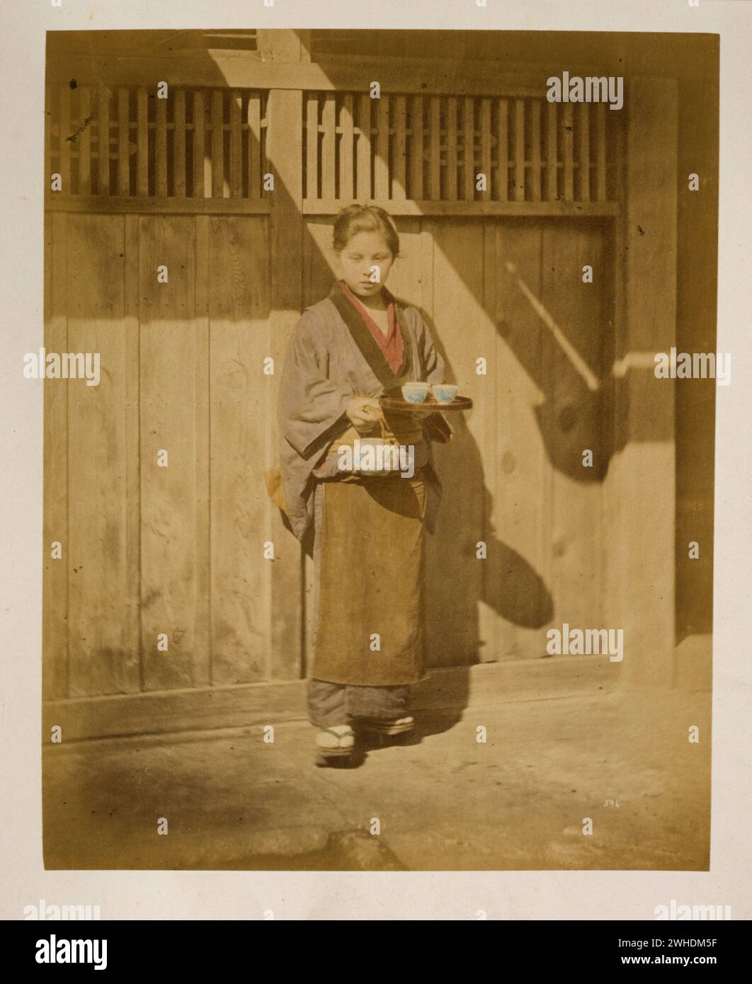 Das Foto zeigt eine junge Frau in voller Länge, stehend, nach vorne gerichtet, mit einer Teekanne und einem kleinen Tablett mit Teetassen; sie trägt vielleicht eine Schürze um ihre Taille und ist möglicherweise eine Kellnerin in einer Teestube. Außenansicht des Gebäudes im Hintergrund... Japan handkoloriert mit Aquarell-Fotografie um 1870er Jahre Stockfoto