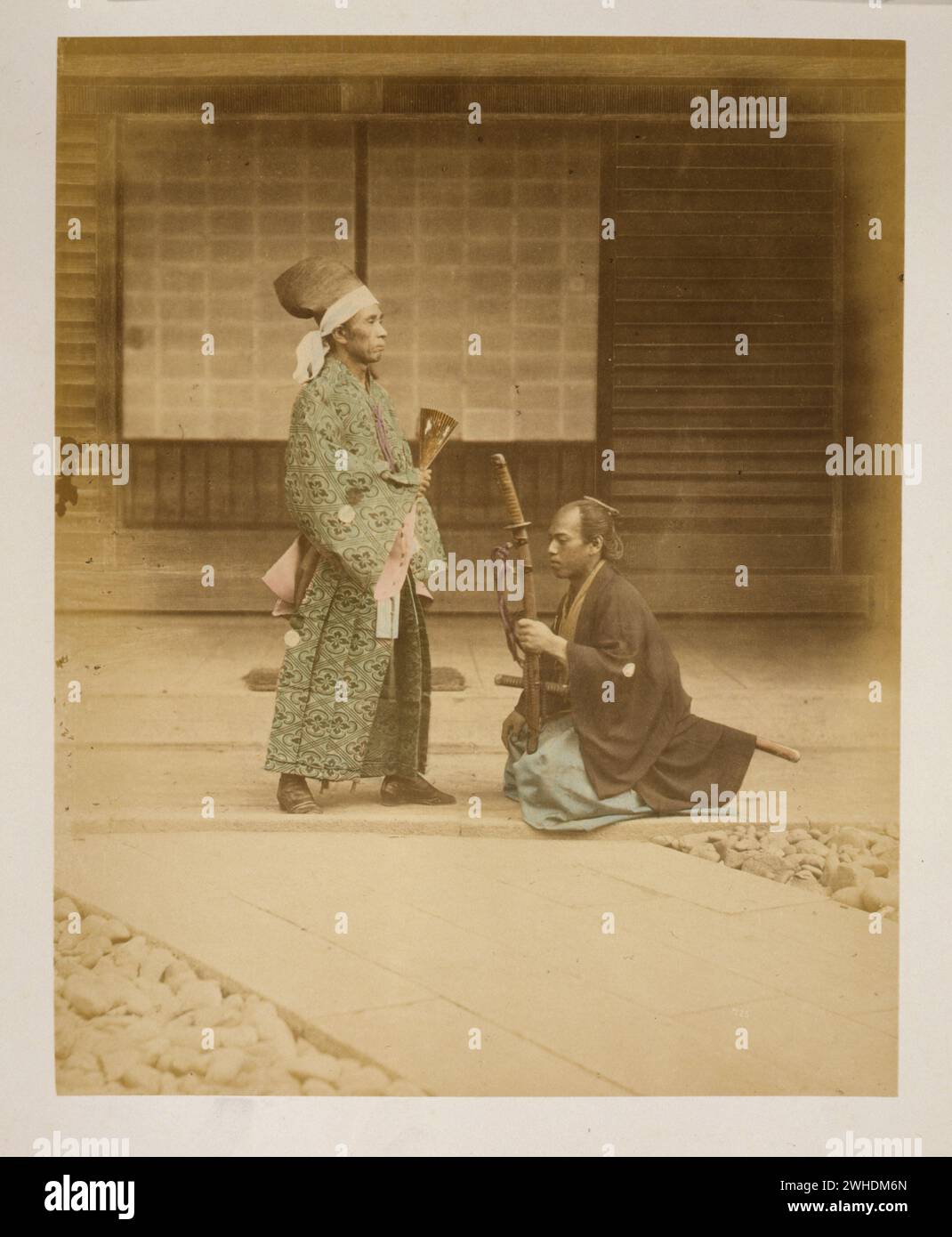 Samurai oder Gefangener kniend vor Regierungsbeamten oder Daimyo- Foto zeigt einen Mann in voller Länge, stehend, nach rechts, und einen Mann, der ein Schwert trägt, kniend vor ihm, nach links gerichtet, hält ein anderes Schwert, als ob er es dem Mann vor ihm anbietet. Außenansicht des Gebäudes im Hintergrund... Japan handkoloriert mit Aquarell-Fotografie um 1870er Jahre Stockfoto
