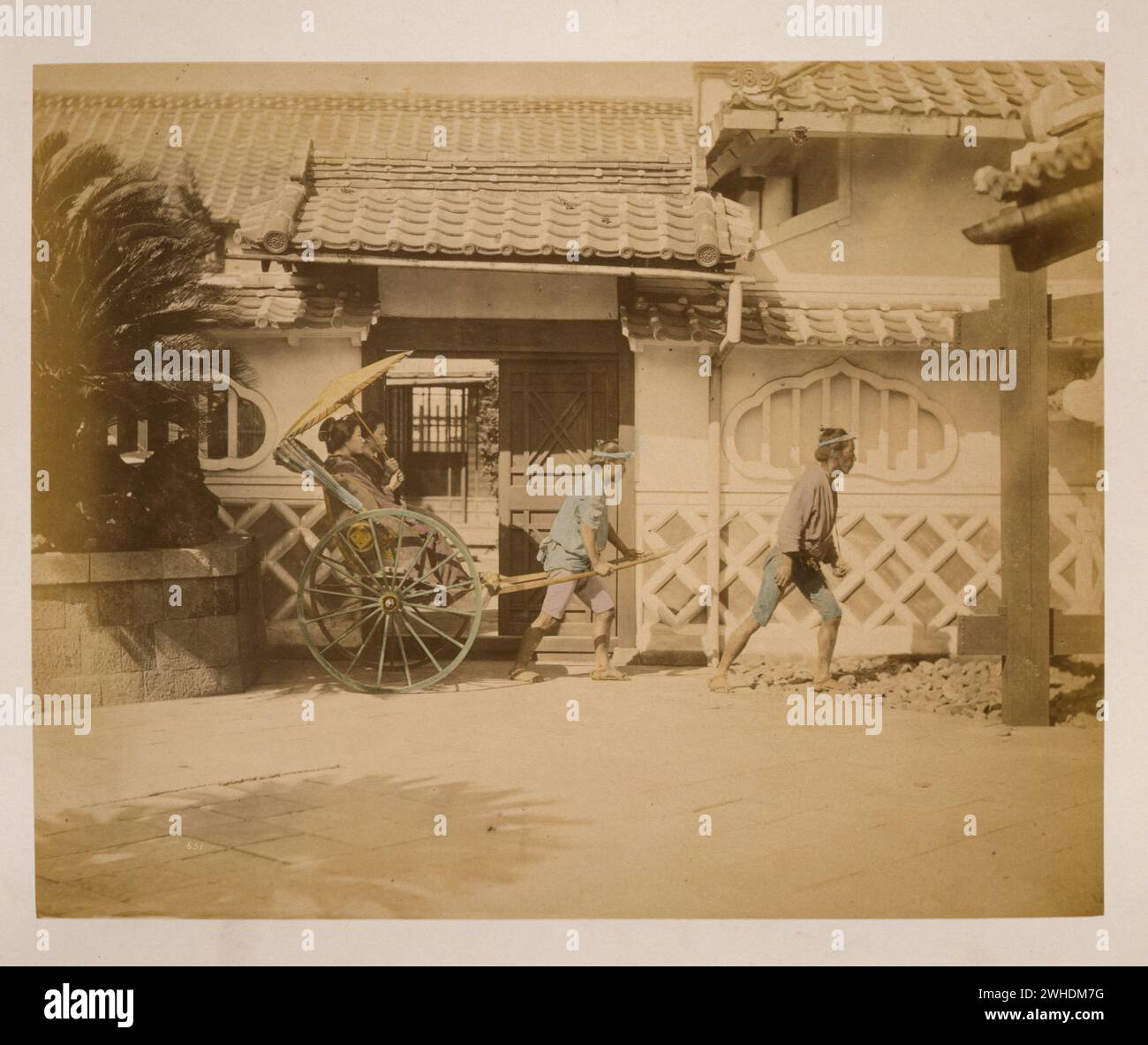 Das Foto zeigt zwei Frauen mit einem großen Sonnenschirm, die in einer Rikscha reiten, gezogen von einem Mann mit einem zweiten Mann, der in der Nähe läuft. Außenansicht des Gebäudes mit gekacheltem Dach im Hintergrund... Japan handkoloriert mit Aquarell-Fotografie aus den 1870er Jahren Stockfoto