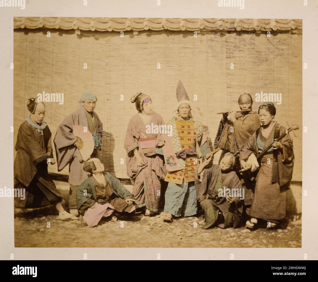 Gruppe von Straßenmusikern und Schauspielern vor einem Gebäude mit Schilf- oder Bambusjalousien....Japan handkoloriert mit Aquarellbild um 1870er Jahre Stockfoto